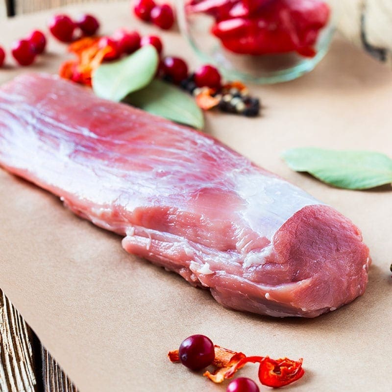 Raw pork tenderloin on brown paper with cranberries, bay leaves, and red peppers nearby.
