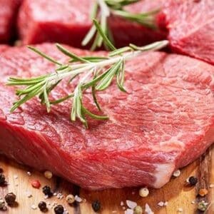 Raw red meat with rosemary and peppercorns on a wooden surface.