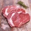 Two raw ribeye steaks on wood with salt grains and herbs in the background.