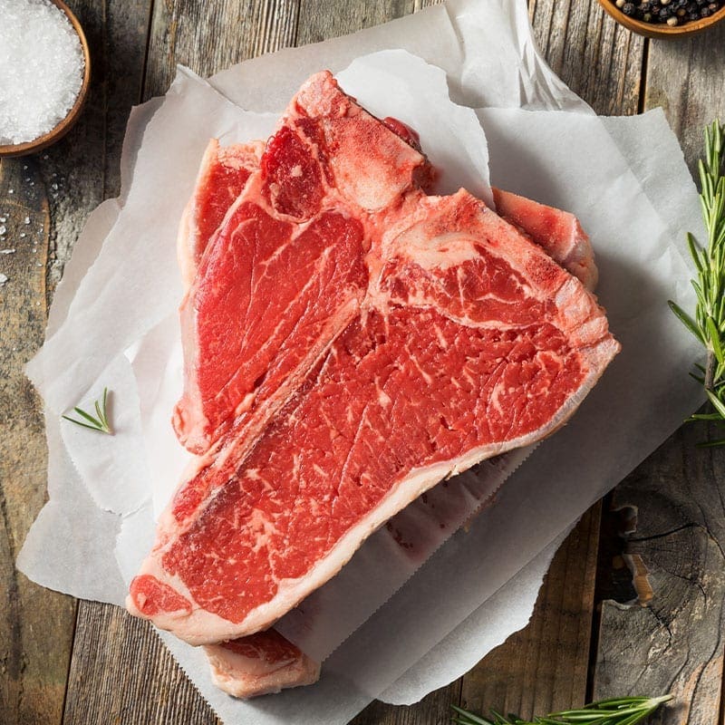 Raw T-bone steak on parchment with rosemary, coarse salt, and peppercorns on a wooden surface.