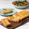Rectangular quiche on board, two slices cut, one on a blue plate, salad in background.