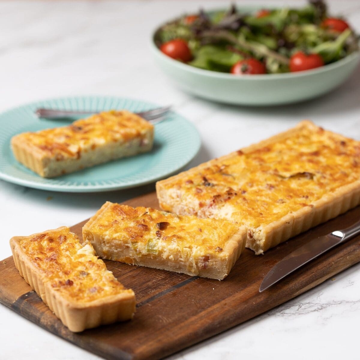 Rectangular quiche on board, two slices cut, one on a blue plate, salad in background.