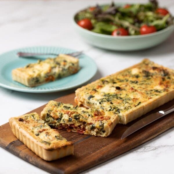 Rectangular vegetable quiche on board, sliced with piece on blue plate near salad bowl.