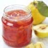Jar of red fruit jam with whole and halved yellow fruits on light surface in background.