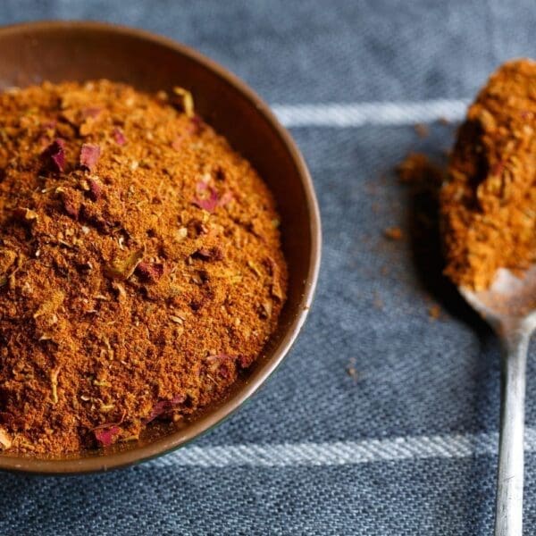 Bowl of reddish-brown spice mix with spoon on dark textured fabric background.