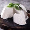 Wedge of ricotta on wooden board with a slice cut, knife nearby, basil garnish, grey background.