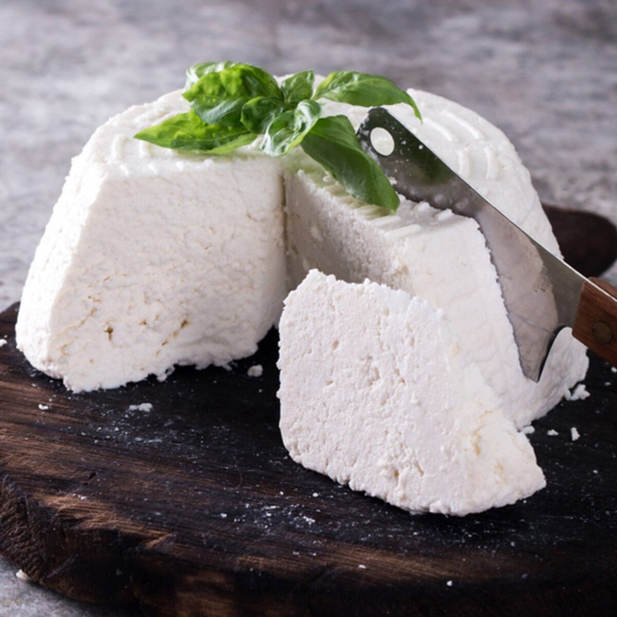 Wedge of ricotta on wooden board with a slice cut, knife nearby, basil garnish, grey background.