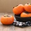 Ripe orange persimmons on a wooden surface, some in a basket, one on a checkered cloth.