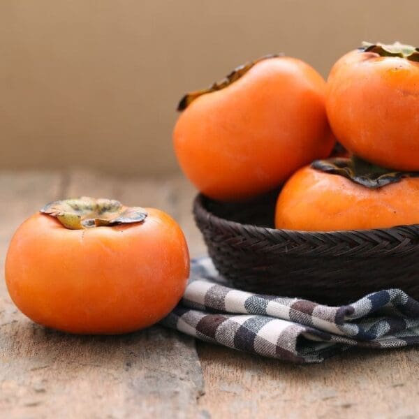 Ripe orange persimmons on a wooden surface, some in a basket, one on a checkered cloth.