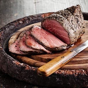 Roast beef on wooden board, sliced to show juicy pink center, with carving knife beside.