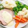 Plate with roast meat, creamy sauce, mashed potatoes, broccoli, cauliflower, carrot sticks.