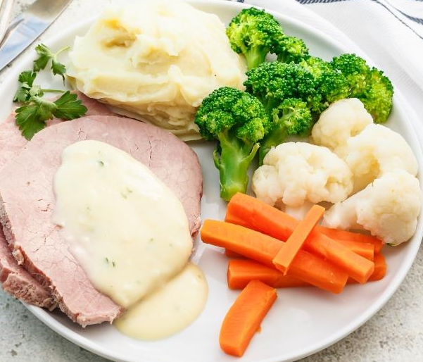 Plate with roast meat, creamy sauce, mashed potatoes, broccoli, cauliflower, carrot sticks.