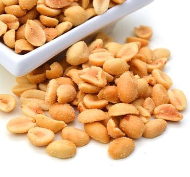 Roasted peanuts spilling from a white dish onto a white surface, lightly salted.