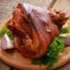 Roasted pork knuckle on wooden platter with parsley and purple onion wedges.