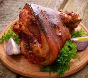 Roasted pork knuckle on wooden platter with parsley and purple onion wedges.