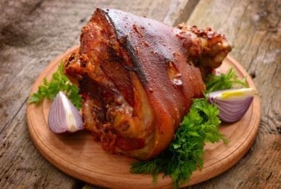 Roasted pork knuckle on wooden platter with parsley and purple onion wedges.