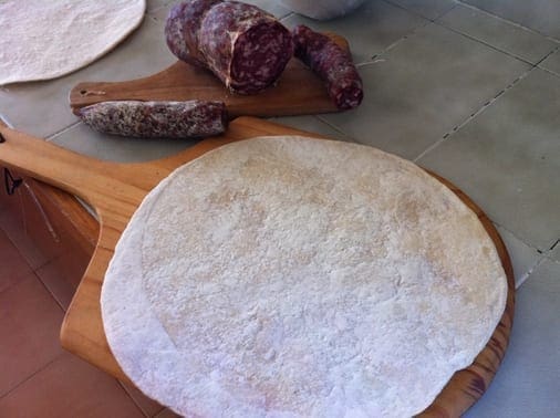 Round dough on wooden pizza peel with cured meat pieces on tiled surface.