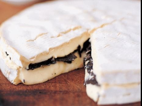 Round cheese wheel with white rind and truffle layers, on wooden surface, portion cut.