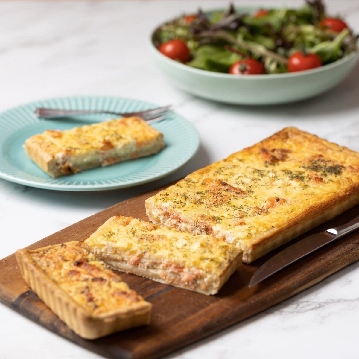 Savory rectangular tart on a wooden board with salad and tart slice on aqua plate nearby.