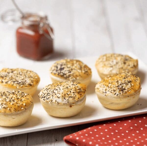 Six seeded pastries on a white plate, red sauce jar, and dotted napkin on wooden surface.