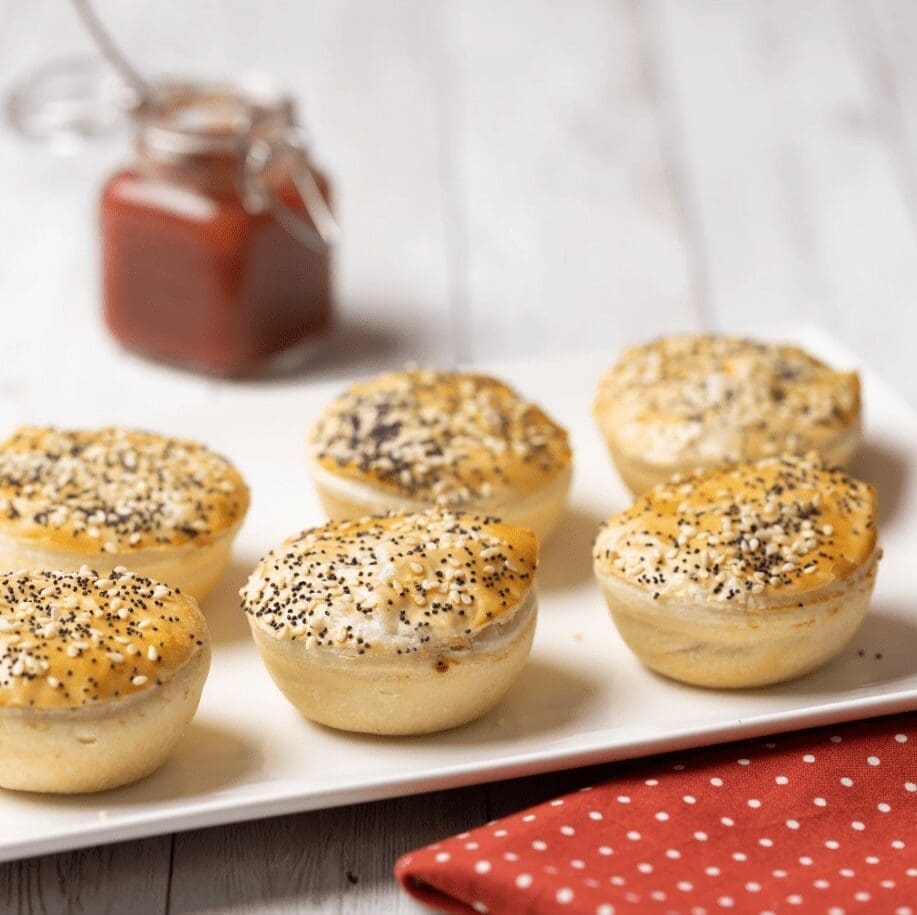 Six seeded pastries on a white plate, red sauce jar, and dotted napkin on wooden surface.