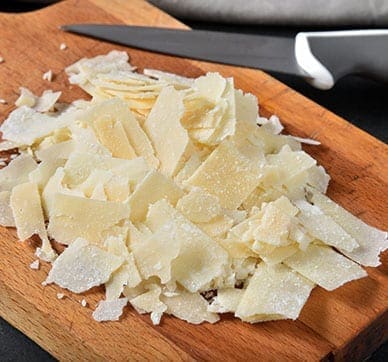 Shaved cheese on wooden board with knife nearby.