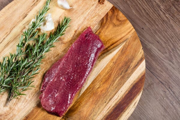 Raw steak on wooden board with rosemary and garlic clove nearby.