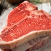 Raw T-bone steak on parchment with rosemary, coarse salt, and peppercorns on a wooden surface.