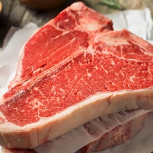Raw T-bone steak on parchment with rosemary, coarse salt, and peppercorns on a wooden surface.