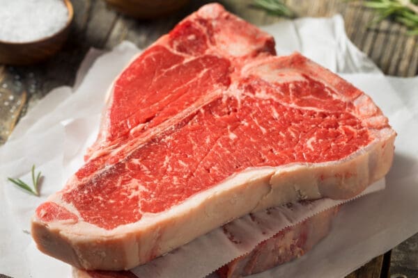 Raw T-bone steak on parchment with rosemary, coarse salt, and peppercorns on a wooden surface.