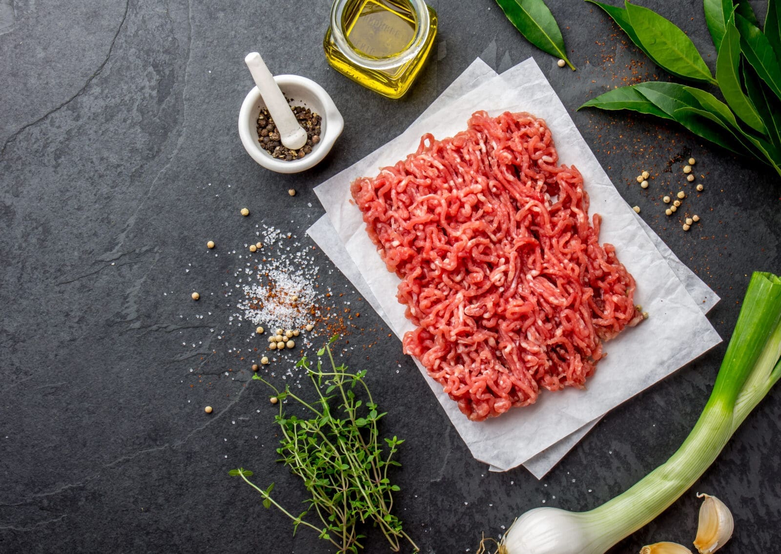 Raw ground meat on parchment paper with herbs, pepper, olive oil, salt, and bay leaves.
