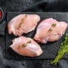 Two raw chicken thighs on a wooden board with a sprig of fresh herbs.