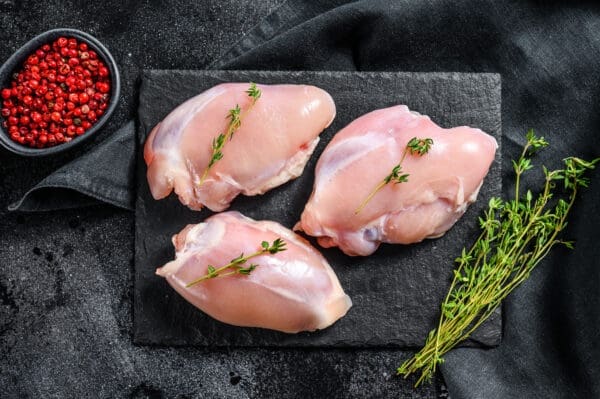 Two raw chicken thighs on a wooden board with a sprig of fresh herbs.