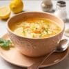 Bowl of vegetable soup with lemon, parsley, spoon; salt, pepper, halved lemon, napkin nearby.