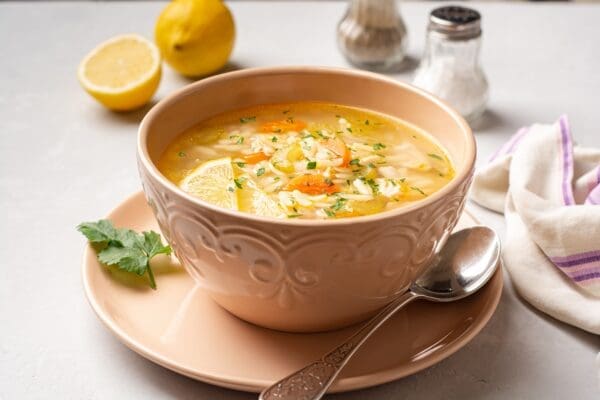 Bowl of vegetable soup with lemon, parsley, spoon; salt, pepper, halved lemon, napkin nearby.