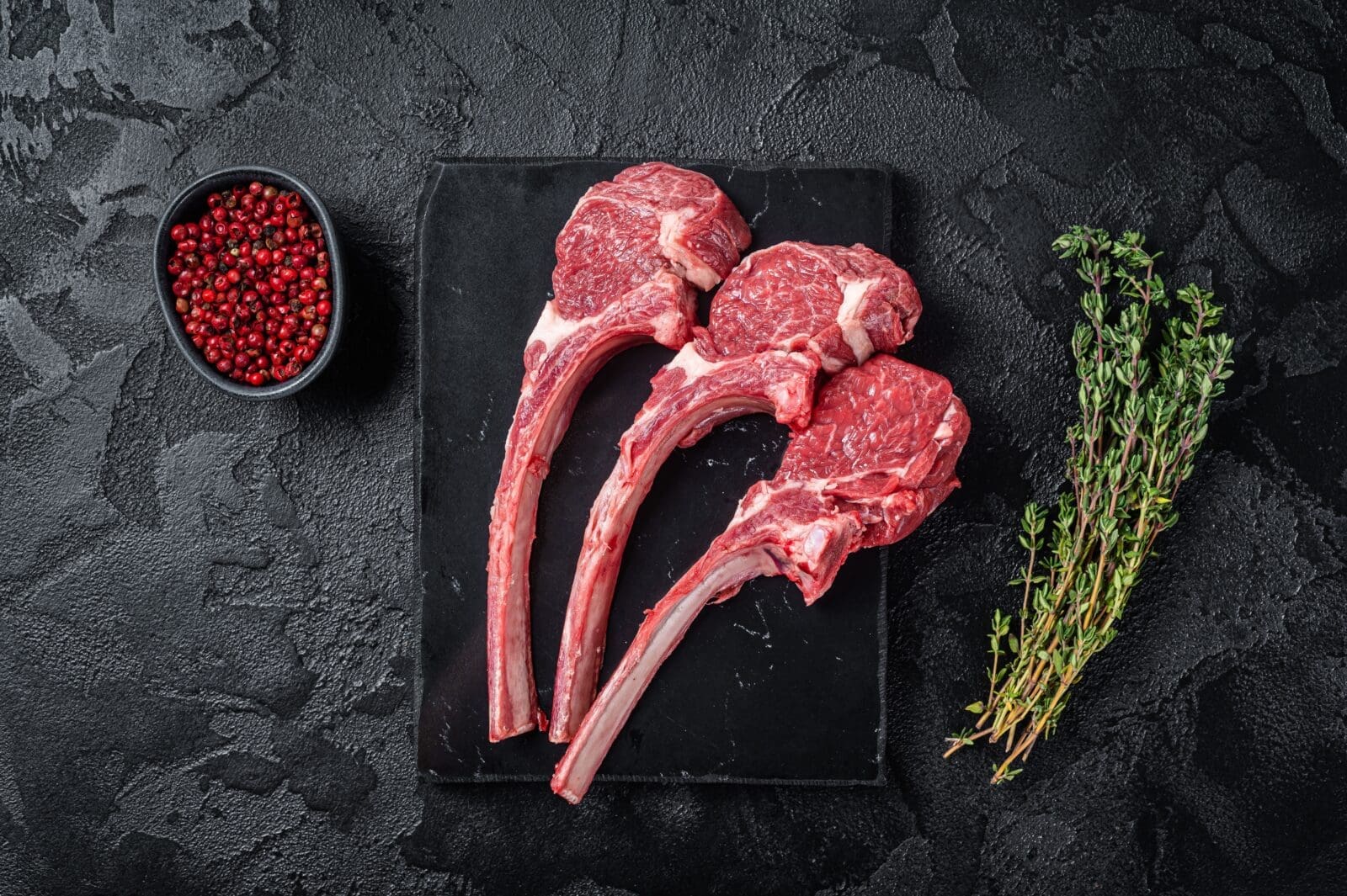 Three raw lamb chops, red peppercorns, and thyme on a black slate surface.