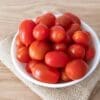 White bowl with small red tomatoes on burlap and wooden surface.