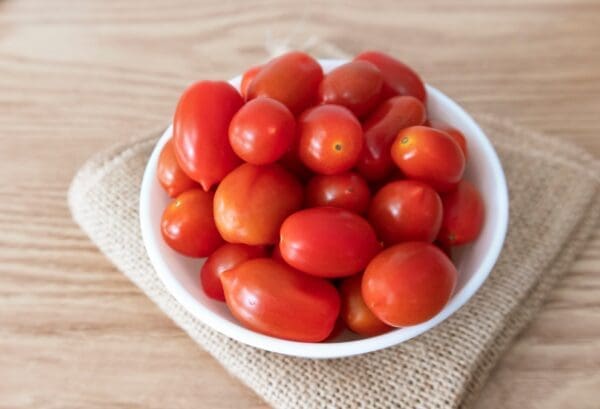 White bowl with small red tomatoes on burlap and wooden surface.