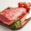 Raw beef steak on a cutting board with knife, parsley, salt, tomatoes.
