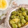 Decorative silver dish with green olives, lemon zest, garlic, thyme; squeezed lemon nearby.