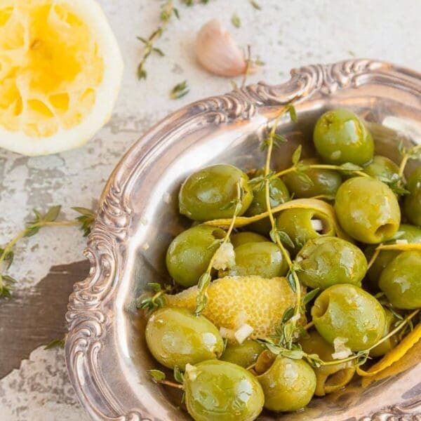 Decorative silver dish with green olives, lemon zest, garlic, thyme; squeezed lemon nearby.
