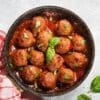 Skillet of meatballs in tomato sauce with basil, red-checkered cloth beside, basil leaves nearby.