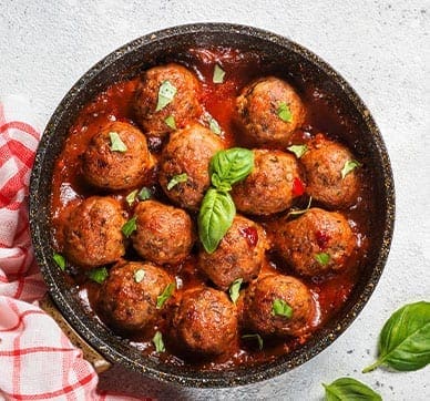 Skillet of meatballs in tomato sauce with basil, red-checkered cloth beside, basil leaves nearby.
