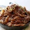 Sliced beef in glossy sauce with sesame seeds and green onions in a dark bowl.