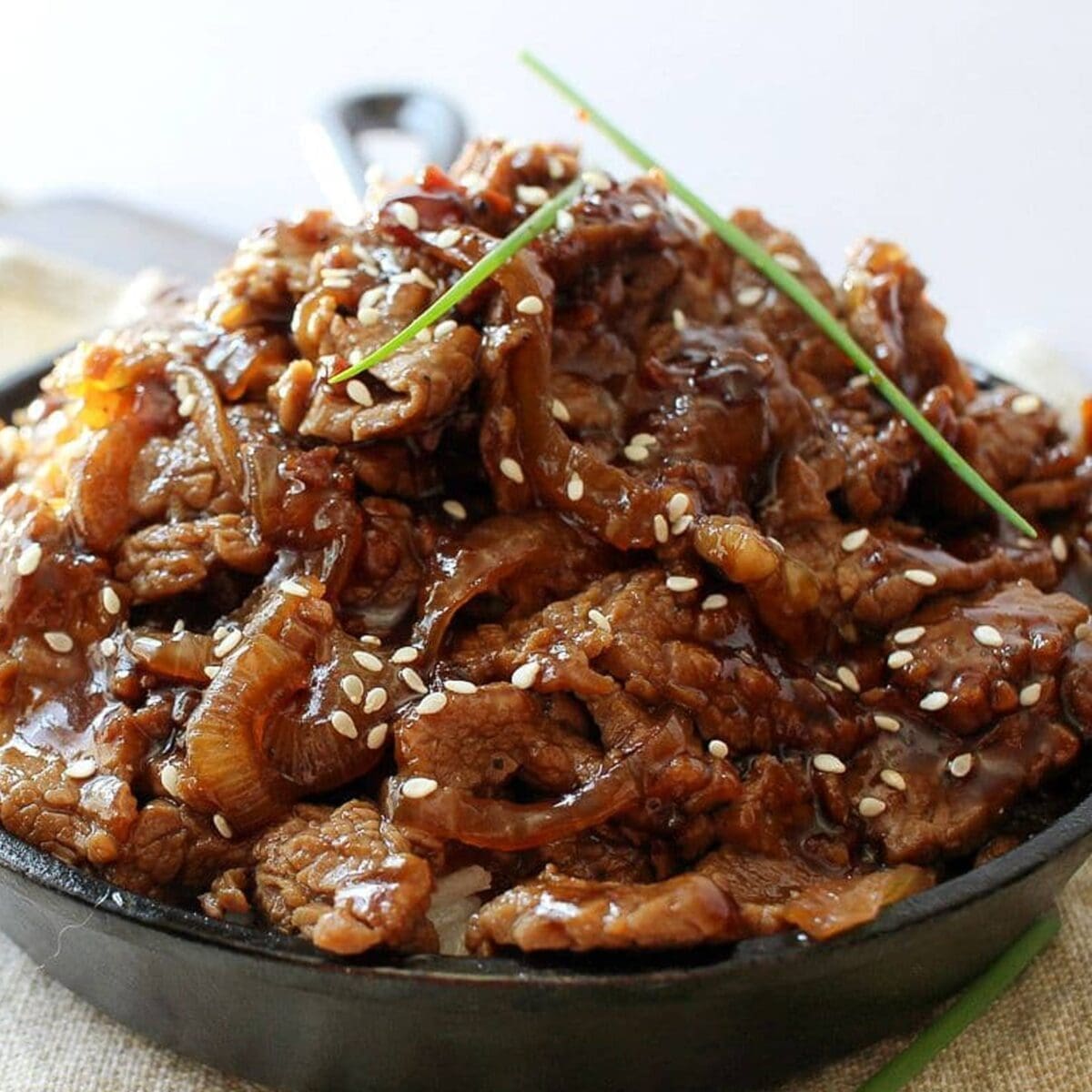 Sliced beef in glossy sauce with sesame seeds and green onions in a dark bowl.
