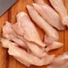 Sliced raw chicken breast on a wooden cutting board with a knife nearby.