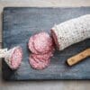 Partially sliced salami wrapped in netting with round slices and knife on a wooden board.