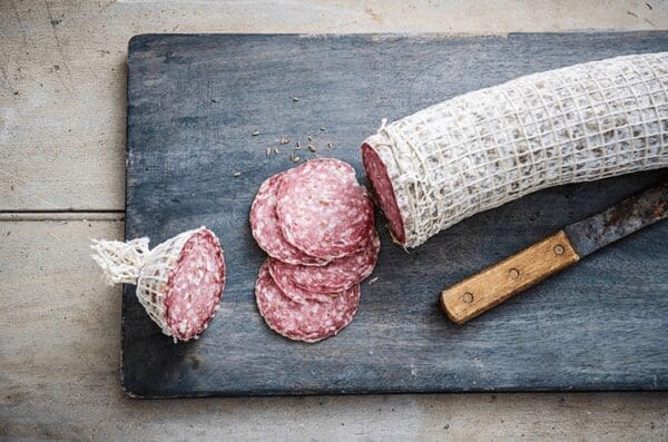 Partially sliced salami wrapped in netting with round slices and knife on a wooden board.