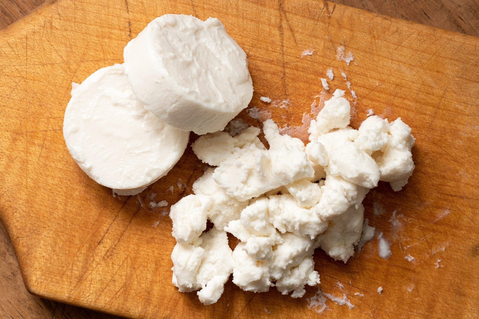 Sliced white cheese and crumbles on a wooden cutting board.