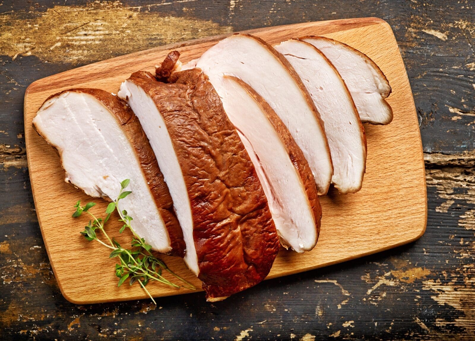 Smoked turkey slices on wooden board with herbs, set on rustic surface.
